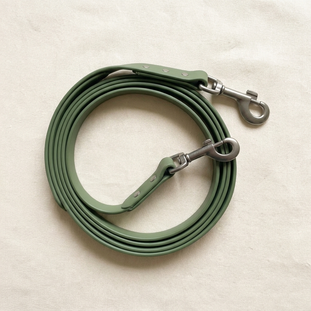A moss-green coated waterproof dog lead with matte trigger clip, coiled on cream linen